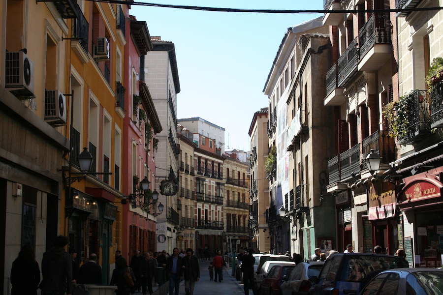La Latina, Madrid – narrow historic street with colorful facades, wrought-iron balconies, shops, pedestrians, and parked cars on a sunny day.