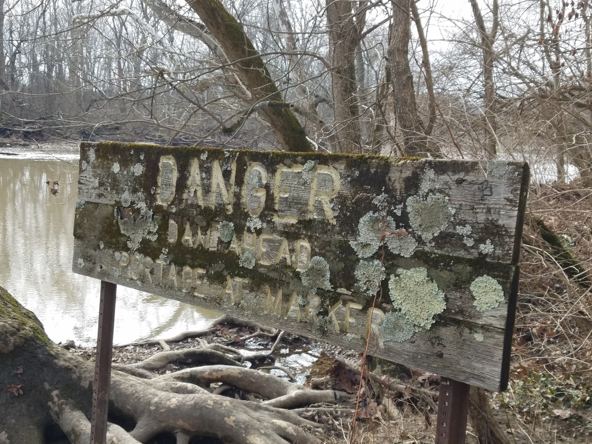 An old wooden sign warning about the lowhead dam just down stream. There are newer looking metal signs. The lichen encrusted sign reads "Danger, Dam Ahead. Portage at Marker". This can be found east of Antrim lake on the western shore of the river, along the trails that snake through the wooded area of the park. #urbanjungle
