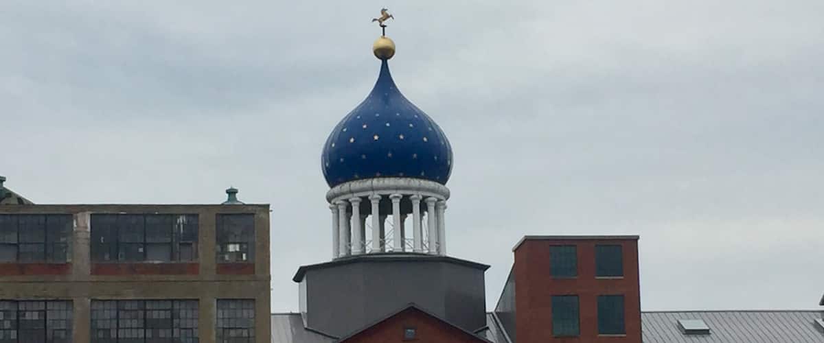 Underneath that blue dome lies the east armory of a factory once owned by a man who changed American history, the innovator of the revolver, Samuel Colt. Sprawling around the crumbling red brickwork of the old Colt’s Patent Firearms Manufacturing Company, once the largest private armory in the world, are the remnants of Coltsville, the utopian village he built for his workers. 
A model of 19th century industrial paternalism, Coltsville included a church, a social hall for dances and lectures, workers’ housing, a giant landscaped park home to deer and peacocks, sculpted botanical gardens, and thousands of feet of greenhouses filled with tropical fruit and flowers. Samuel Colt felt such responsibility for the welfare of his workers outside the factory that he went so far as to build Potsdam Village, to replicate the feel of the German village from which he imported his skilled craftsmen to work in his willow ware factory.
The National Park Service began having on-site presence in 2015, with workshops and tours, including up into the Blue Onion Dome which affords magnificent views of downtown Hartford, the Connecticut River and Colt Park, formerly the back yard of Armsmear, Samuel & Elizabeth Colt’s mansion. By 2016, an NPS ranger had taken up residence in the South Armory and was giving walking tours of Coltsville.
The old South, East and North armories are under renovation and the two 1855 brownstone buildings, the oldest structures on site, will become the National Park Service Visitor Center. The armories themselves have been or are being renovated into apartments, offices and education facilities.
