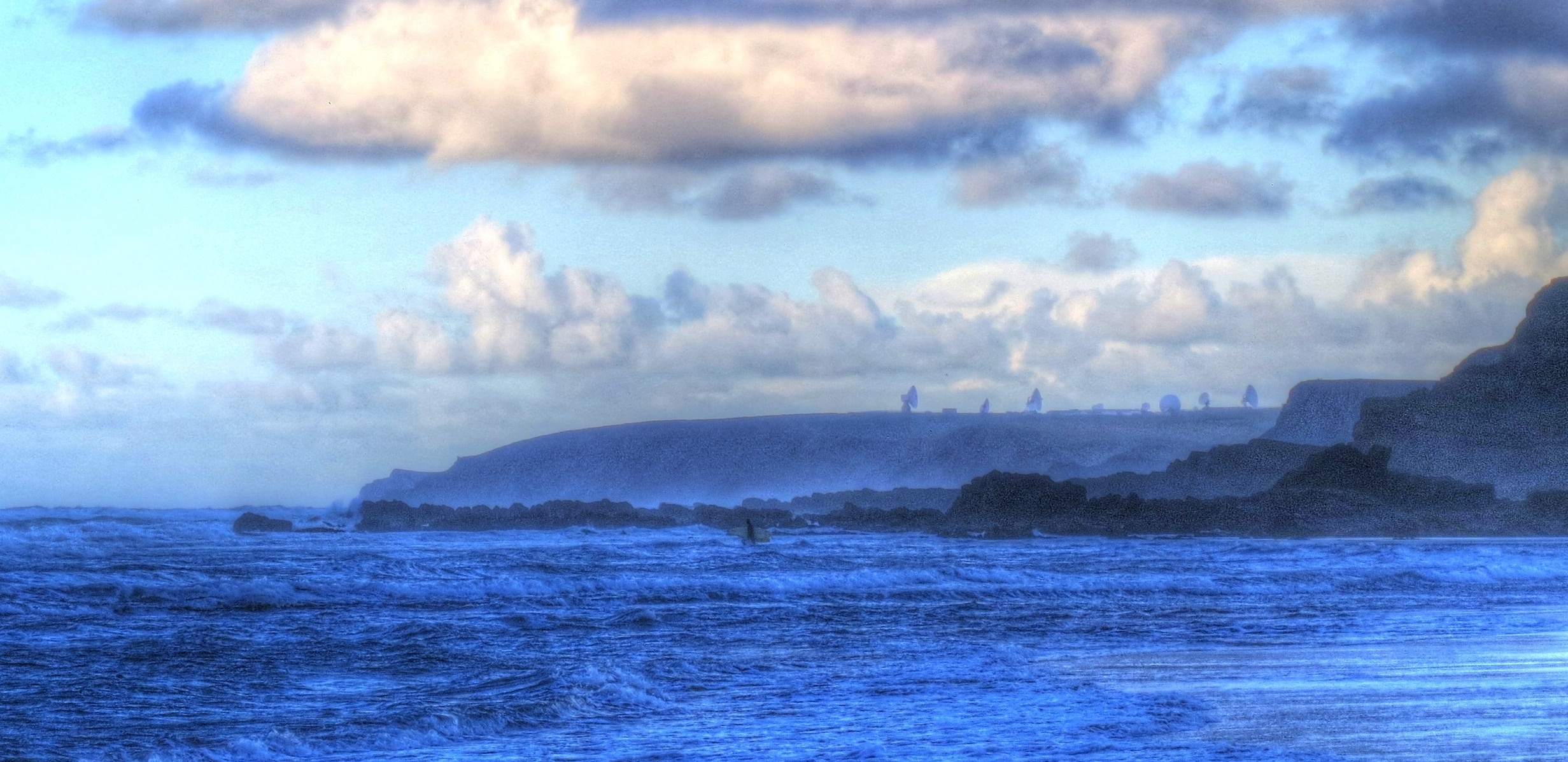 This photograph was taken from Widemouth Bay beach, showing a surfer going into the see there. In the background can be seen the dishes of GCHQ Bude.