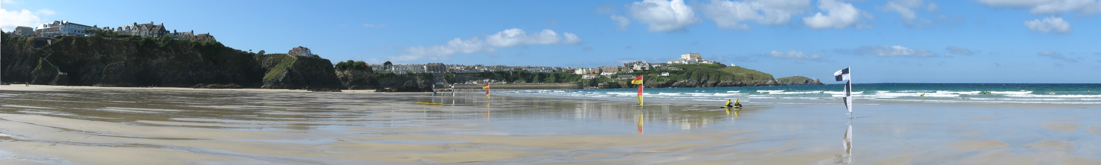 Panorama westward from Tolcarne Beach, Newquay, at close to low-tide. From left to right: Newquay buildings on clifftop; Newquay harbour entrance; The Atlantic Hotel and headland; Towan Head in distance. Two lifeguards sitting on boards in the foreground.