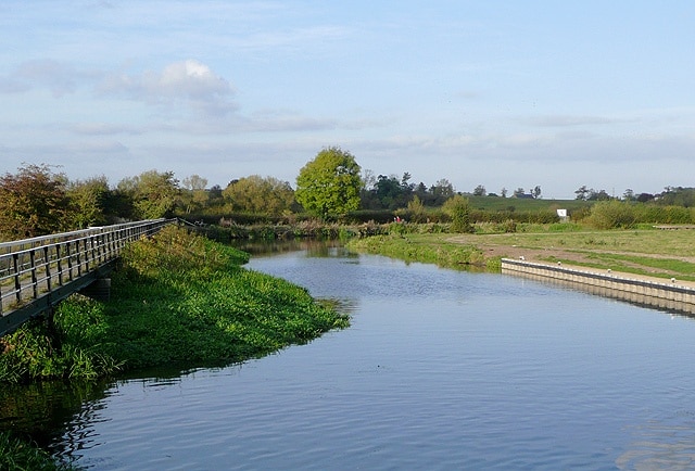 River Trent at Alrewas, Staffordshire. Seen from Alrewas lock on the Trent and Mersey Canal. The walkway on the left is the towpath bridge across the river, which is entering from the left. The metal piling on the right allows firm mooring for narrowboats waiting to go into Alrewas Lock and into the canal. About three hundred metres round the bend to the right, a large weir allows the canal to continue to the north of the river, which continues to the east. Narrowboats must not be taken onto this river section when it is in flood.