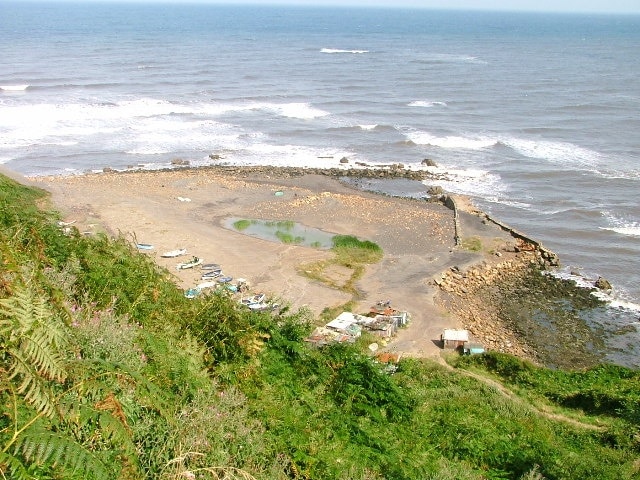 Port Mulgrave Harbour. An almost identical picture to the other undated one showing the recent filling of the harbour. I suspect this is deliberate to prevent erosion of what remains of the harbour. There now is a pond with bullrushes in. Port Mulgrave owes its existence to the ironstone industry. The blocked up mine entrance can still be seen 50' above high water above what remains of the harbour. Tunnelling began in 1854 and work on the harbour had started two years later. By the 1870's new more productive seams were found three miles away at the secluded valley of Easington Beck in Grinkle Park. The only feasible method of transporting the stone out was by sea and so the original tunnel at Port Mulgrave was extended for a further mile to connect to the Grinkle Park mine. Gradually the Port Mulgrave mine itself was abandoned but the harbour continued to be used for Grinkle Park ore until 1917 when a connection was made to the Middlesbrough to Whitby railway owing to the wartime dangers to shipping. The machinery at the harbour was finally dismantled in 1934 during which the wooden gantry accidentally caught fire. Later the Royal Engineers destroyed the breakwater to prevent German forces using the place for an invasion.