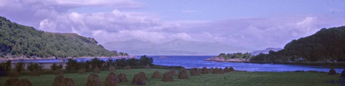 Photo "Loch Feochan, Argyll & Bute taken 1963 Looking over farmland towards the Isle of Mull, which may possibly be seen in the distance. Note the interesting hay stacks. The land at right is Ardentallen Point. The rock in the middle of the channel might be Sgeir Dhubh." by Christine Matthews (Creative Commons Attribution-Share Alike 2.0) / Cropped from original