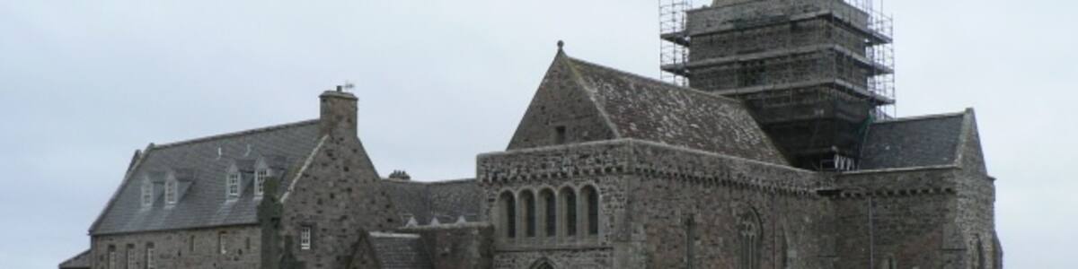 Photo "Isle of Iona: the abbey Iona Abbey has had a long history of erection and destruction. Columba came here and built a wooden monastery in AD563 which was burned down by the Vikings on their second attack, in 802. A replacement was built from 818, this time in stone a little south of the original and where the present-day buildings stand. This was badly damaged by a further Viking raid in 825. Reginald MacDonald of Islay rekindled Iona's religious importance and a large abbey was build in the 1200s with further work in the 1400s. However, after the Reformation in 1560 the building gradually decayed until 1874 when the 8th Duke of Argyll commissioned and architect to preserve the ruins. In 1902 the newly established Iona Cathedral Trust embarked on the Abbey's full restoration and it is largely the results of this comparatively recent work that we see today." by Chris Downer (Creative Commons Attribution-Share Alike 2.0) / Cropped from original