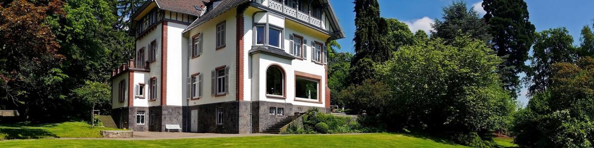 Photo "Die Villa Gail liegt im Gail’schen Park im Biebertaler Ortsteil Rodheim in Hessen. Erbaut wurde die Villa in den Jahren 1896 und 1897 im Auftrag von dem Gießener Tabak-, Zigarren- und Keramikfabrikanten Gail als sommerlichen Landsitz. Rechts ein 1896 gepflanzter Riesenmammutbaum mit 32 m Höhe." by Rainer Lippert (Creative Commons Attribution-Share Alike 3.0) / Cropped from original