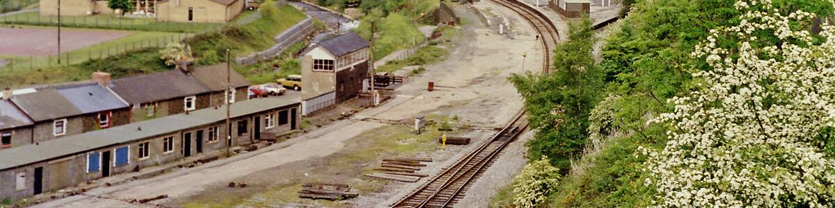 Photo "Junction at Aberbeeg of railways and valleys, 1990. View NW from the A463 road up the Ebbw Valley, towards Ebbw Vale (to the left, Ebbw Fawr), with the railway still open - just about) and to the right up the Ebbw Fach valley to Abertillery, Nantyglo and Brynmawr. In this 1990 view it appears that there are still station platforms for both routes, but the station had been closed for passengers on 30/4/62 and for goods from 28/11/66. The line up the Ebbw Fawr remained open to serve Ebbw Vale Steelworks and a passenger service to Ebbw Vale - but not stopping here - was restored on 6/2/08. The line to the right to Nantyglo and Brynmawr was closed from 30/4/62, although goods to Abertillery continued until 7/4/69, but it still seems to exist here in 1990." by Ben Brooksbank (Creative Commons Attribution-Share Alike 2.0) / Cropped from original
