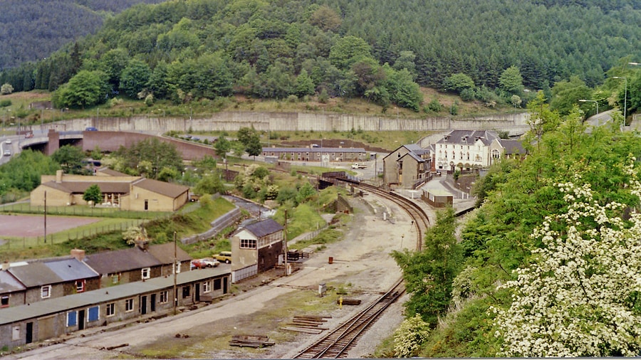 Photo "Junction at Aberbeeg of railways and valleys, 1990. View NW from the A463 road up the Ebbw Valley, towards Ebbw Vale (to the left, Ebbw Fawr), with the railway still open - just about) and to the right up the Ebbw Fach valley to Abertillery, Nantyglo and Brynmawr. In this 1990 view it appears that there are still station platforms for both routes, but the station had been closed for passengers on 30/4/62 and for goods from 28/11/66. The line up the Ebbw Fawr remained open to serve Ebbw Vale Steelworks and a passenger service to Ebbw Vale - but not stopping here - was restored on 6/2/08. The line to the right to Nantyglo and Brynmawr was closed from 30/4/62, although goods to Abertillery continued until 7/4/69, but it still seems to exist here in 1990." by Ben Brooksbank (Creative Commons Attribution-Share Alike 2.0) / Cropped from original