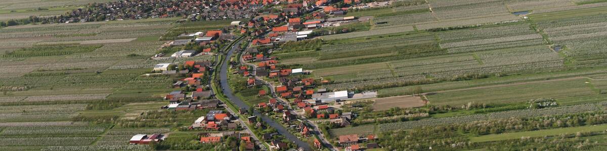 Photo "Luftaufnahme: der kleine Fluss, der das Bild teilt, ist die Lühe, der Ort links ist Steinkirchen, der Ort rechts ist Mittelnkirchen, der Ort vor der Elbe ist Grünendeich, in der Elbe dahinter liegt der Lühesand" by Walter Rademacher (Creative Commons Attribution-Share Alike 3.0) / Cropped from original