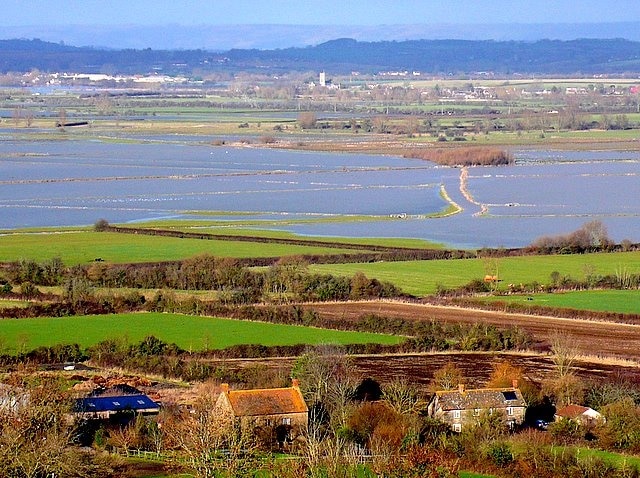 Somerset Levels : Lower Burrow and flooding on West Moor View from the top of Burrow Hill Looking north. The houses in the foreground are at Lower Burrow at the foot of the hill. The flooded fields are on West Moor and the two lines of light coloured reeds mark the line of the main drain of the moor. This drains the moor to the pump house at ST41712353, just out of picture at top left, where the water is pumped up into the River Parret. The river has high embankments and the levels of the river are usually well above that of the moor at this time of the year. The moors are allowed to flood to prevent excessive flood damage to Langport 3km downstream. The houses on the right in the middle distance are at Thorney ST427226 and the church in the centre in the middle distance is at Muchelney Abbey ST429249. The "-ney" ending to a place name on the Levels signifies an island and Thorney and Muchelney are on slightly higher ground that historically saved them from flooding. Langport is in the centre distance and the factories and industrial units on the south side of the town can be seen in the sun in the left distance. The hills in the far distance are the Polden Hills, a low ridge south of, and parallel to the Mendips