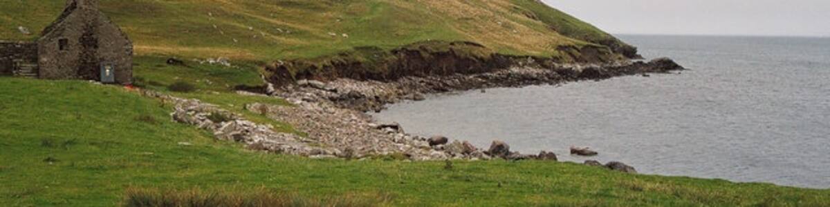 Photo "Beach of East Lunna Voe The photo is taken south of Lunna Kirk. The building appears to be deserted - perhaps an old farm? Erosion of the hillside behind the beach is evident from the exposed face with minor slips." by Richard Kay (Creative Commons Attribution-Share Alike 2.0) / Cropped from original