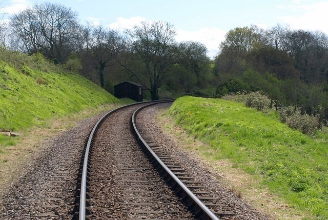 West Somerset Railway. From the same crossing point as 1281019, looking in the other direction down the track towards Combe Florey, past a dark hut rather hidden in shadow compared to the bright grassy banks in the foreground.