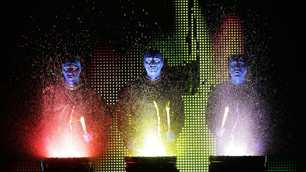 Blue Man Group splashed with paint as they bang on lit up drums onstage in Boston
