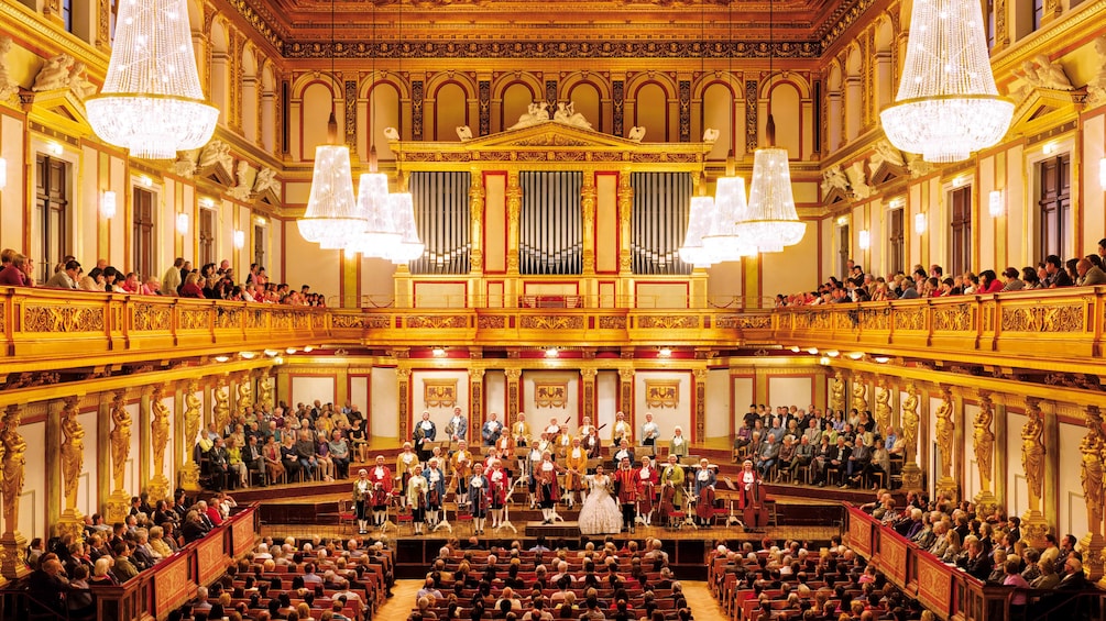 Costumed orchestra and audience in a brightly lit and ornate music hall in Vienna