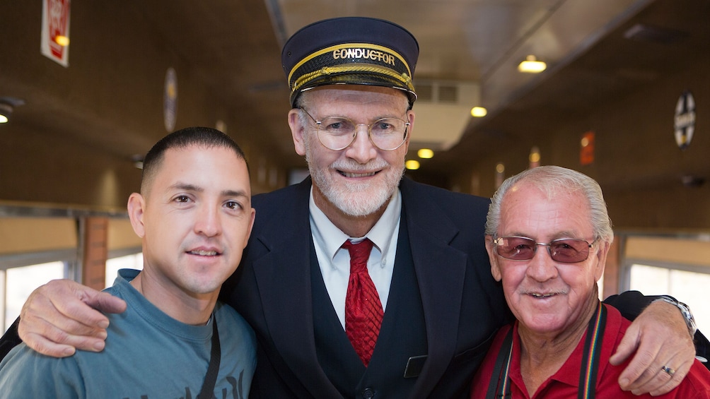 Two men smile with the train conductor aboard the train at the Grand Canyon Railway 