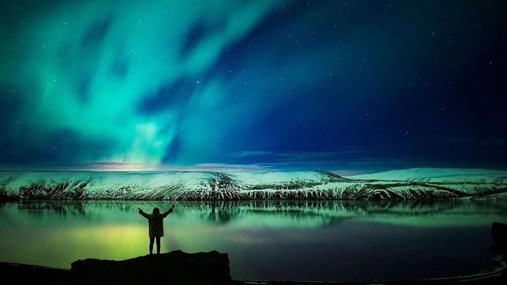 Woman with outstretched arms at edge of lake with Northern Lights above in Reykjavik