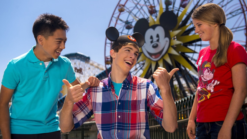 Show item 4 of 10. Young adult wearing Mickey Mouse ears and giving the thumbs up to two friends at Disneyland