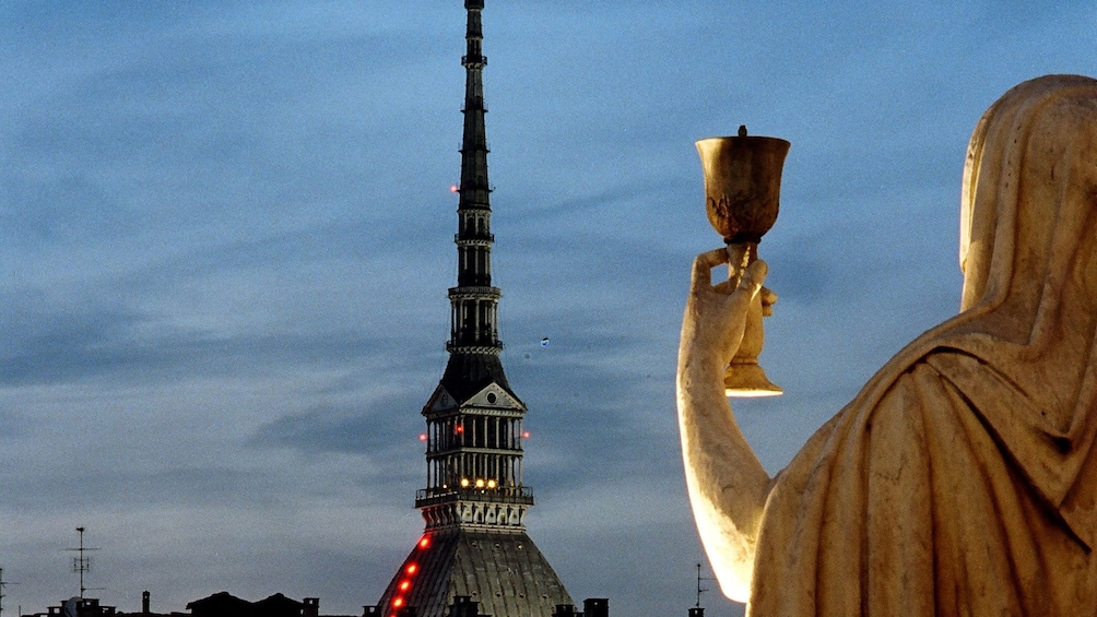 A statue of a female figure holding a cup looking out at a cathedral in Turin