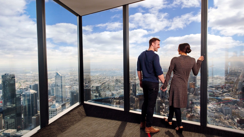 Show item 4 of 7. Couple looking out the large glass windows at the city from the Eureka Skydeck in Melbourne