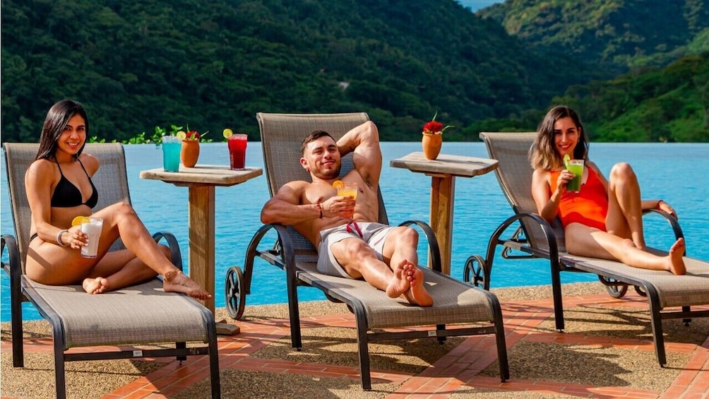 A man and two women lounge by the pool with drinks in hand