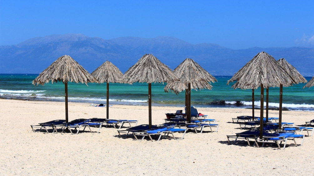 Show item 1 of 10. Chaise lounges under umbrellas along the beach shore with mountains in the distance on Crete Island