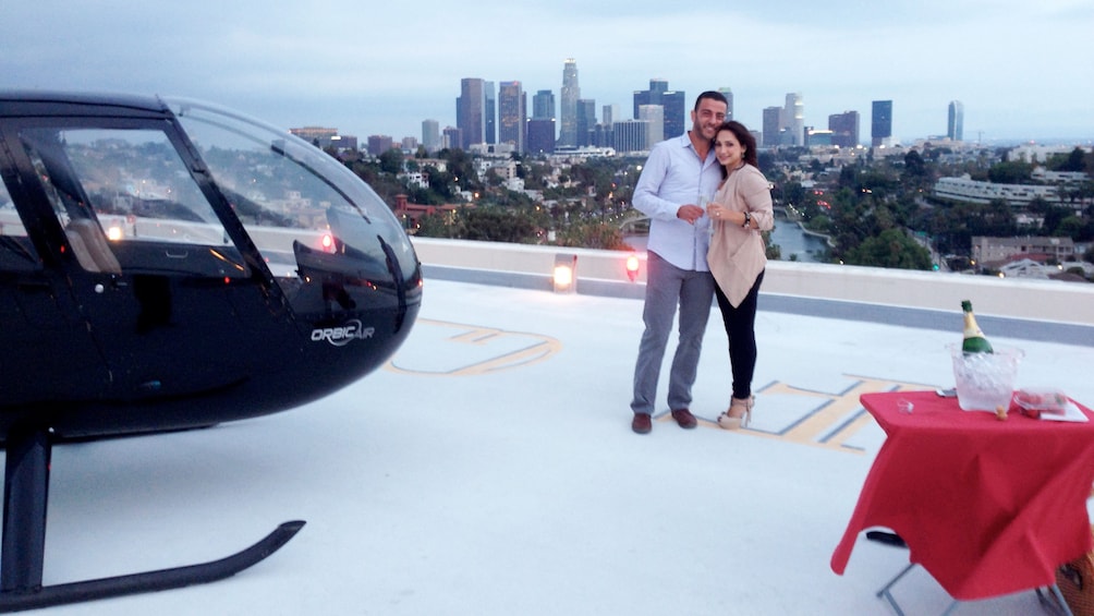 Couple taking a picture overlooking the city with the helicopter off to the left and dinning table off to the right 