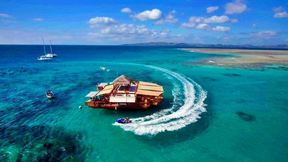 Cloud 9 floating cabana docked in a clear blue sea and a boat spinning around it. 