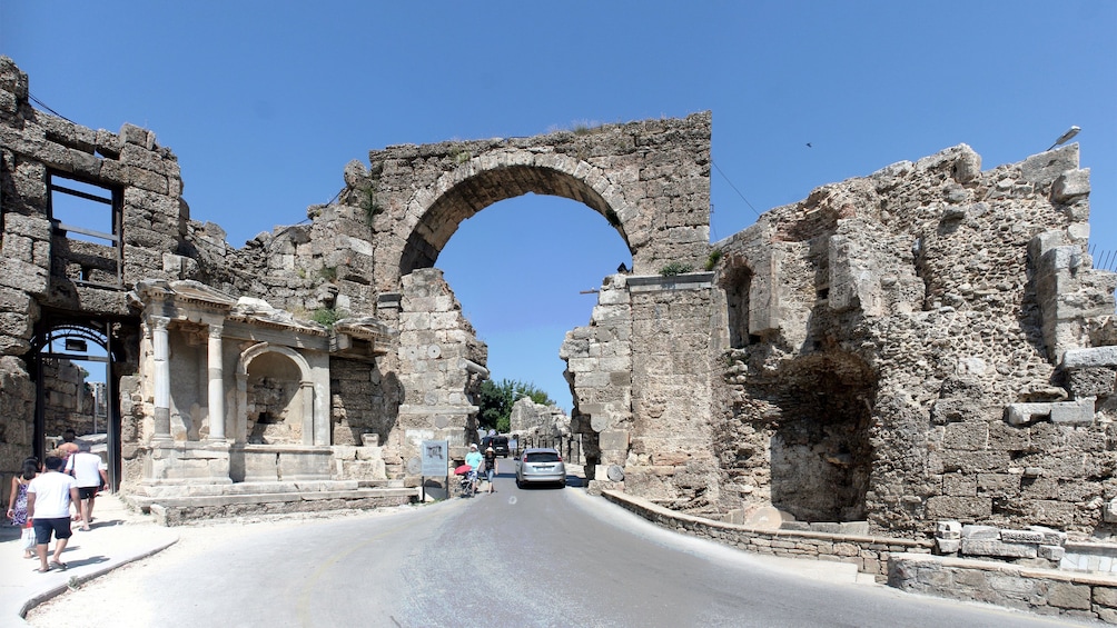 Show item 4 of 5. A car driving under an ancient stone archway at the ruins of Perge