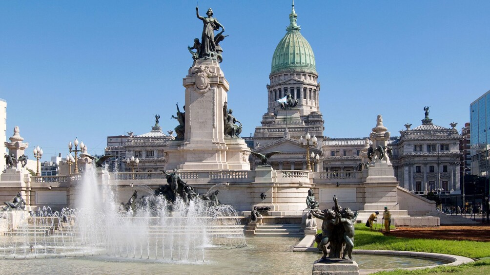 Large fountain with sculptures and a green-domed building in the background