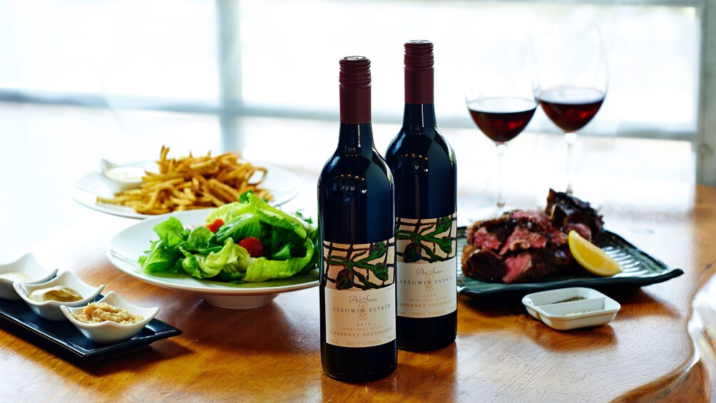 Close up of wooden table with two bottles of red wine, two filled glasses, and food.