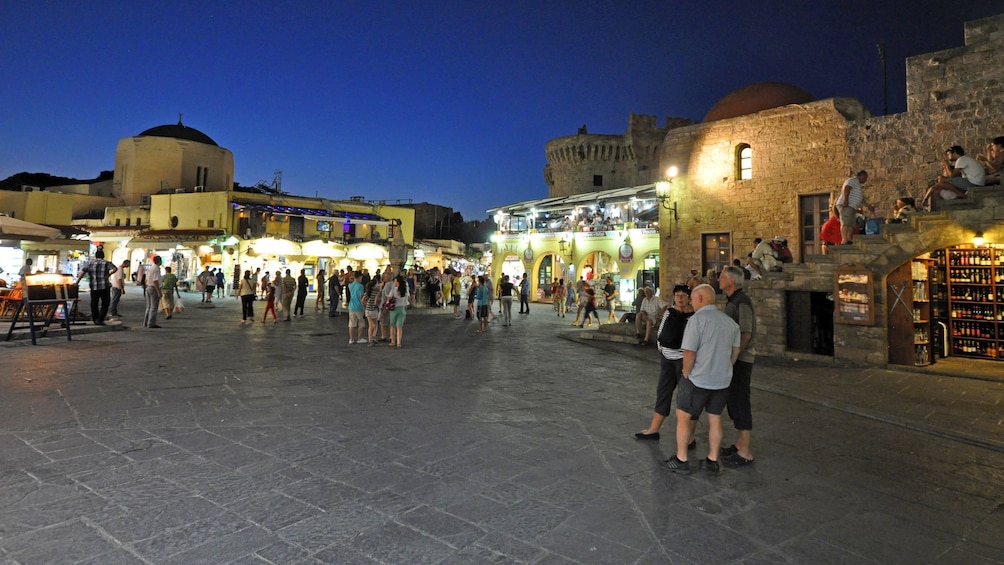 Foto 4 von 5 laden A Grecian plaza with storefronts and patrons at night