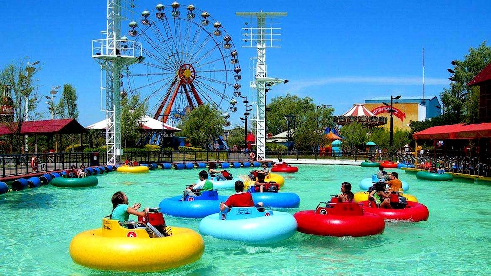 People in multicolored inner tubes with water cannons float in a large pool next to a ferris wheel