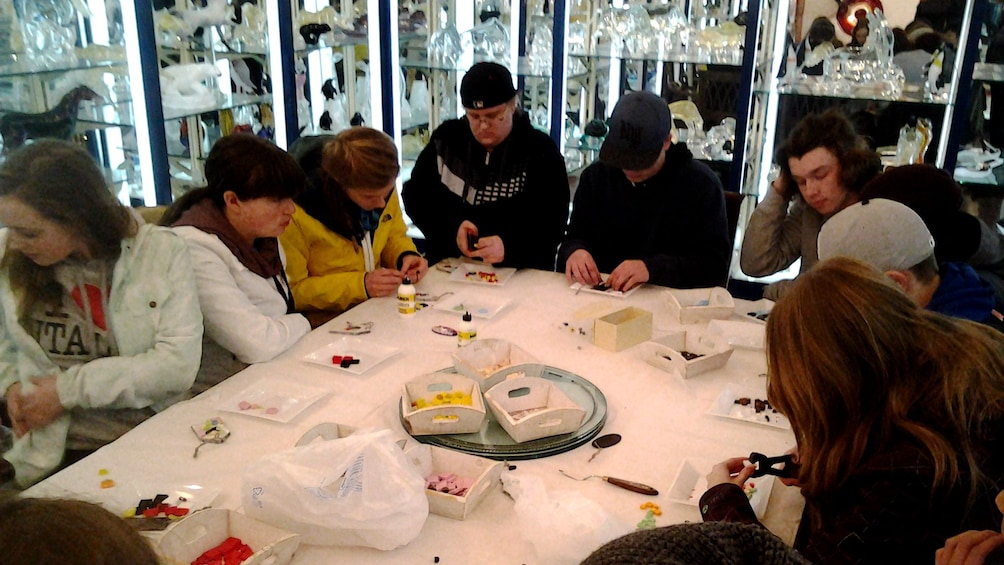 Group of tourists sit around a table and assemble crafts for glass blowing