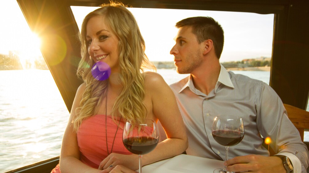Woman smiles to the camera and a man looks out the window while drinking white wine on a wine cruise