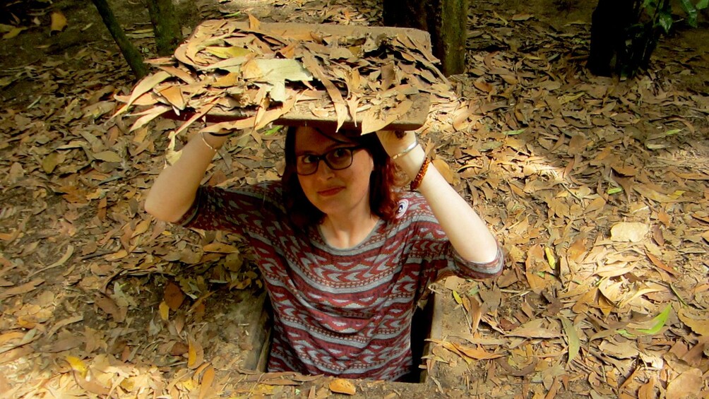 Tourist poses in Ho Chi Minh-Cu Chi tunnels lifting a tunnel lid with leaves on it