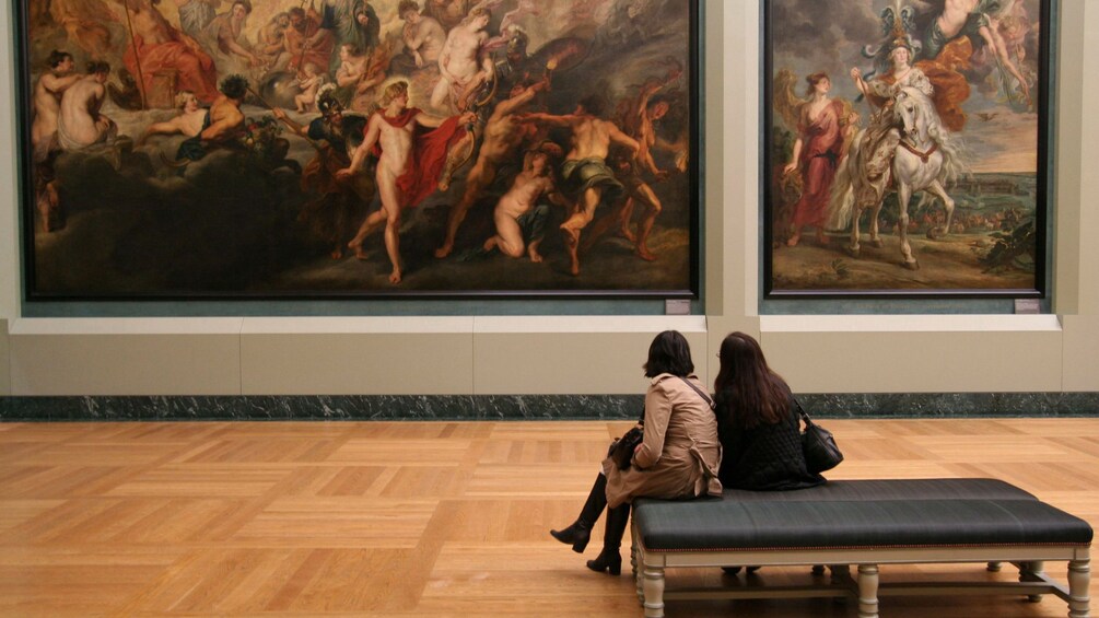 Two people sit on a bench in the Louvre and look at a large painting