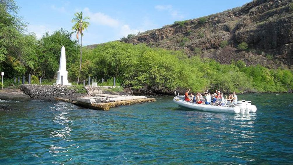 Boat full of people pulling up to a monument on the shore of Hawaii 