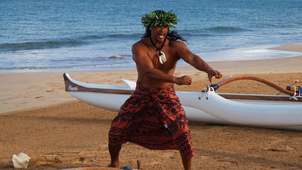 Hawaiian man dances on beach next to an outrigger canoe.
