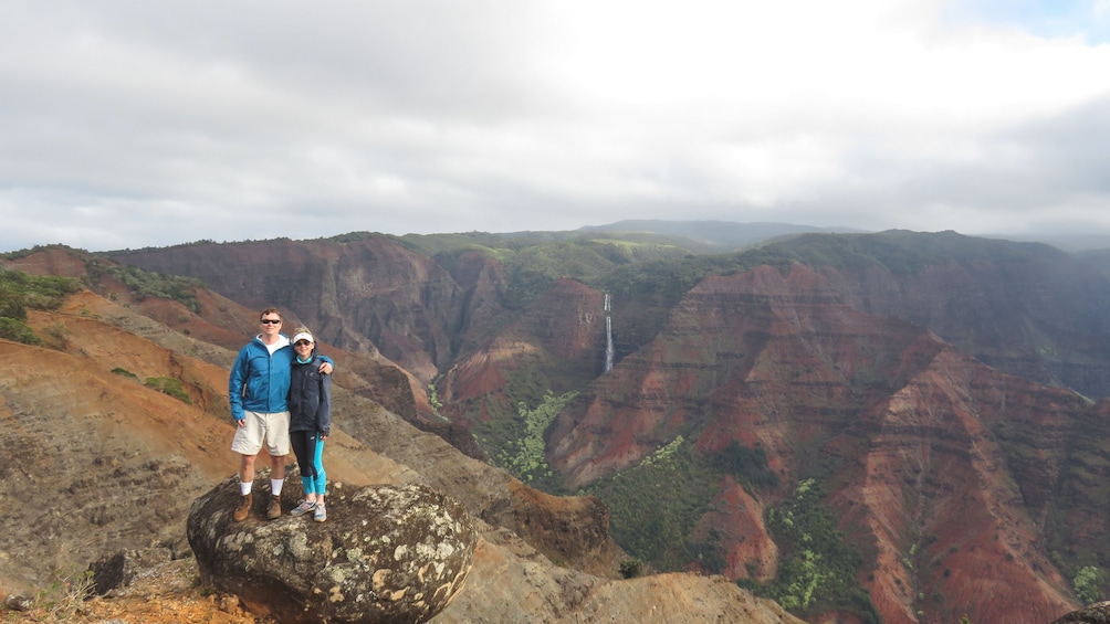 Couple standing together on large rock at mountain peak, with beautiful view of canyons in the distance.