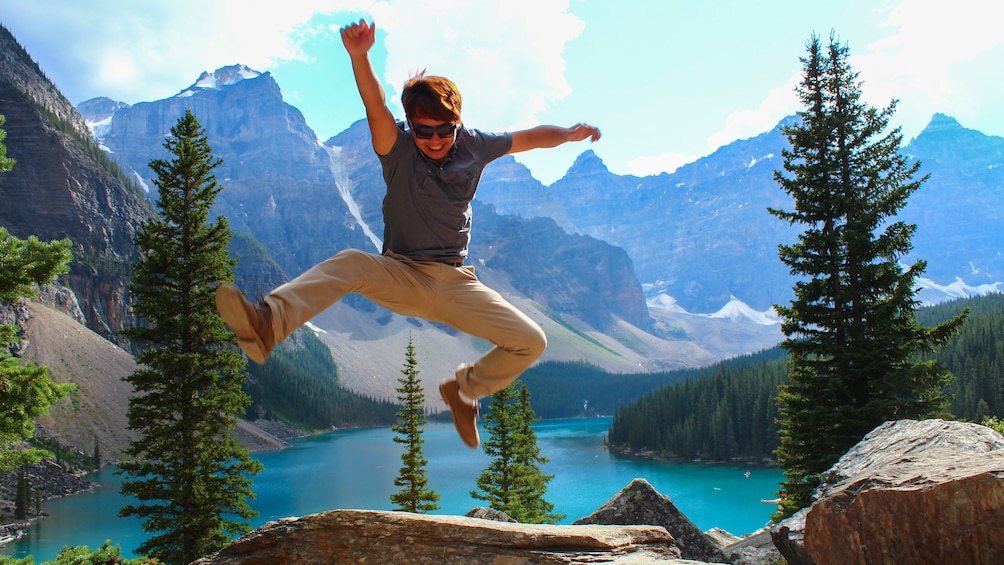 Young man captured mid-air jumping, with beautiful rocky mountain landscape in the distance.