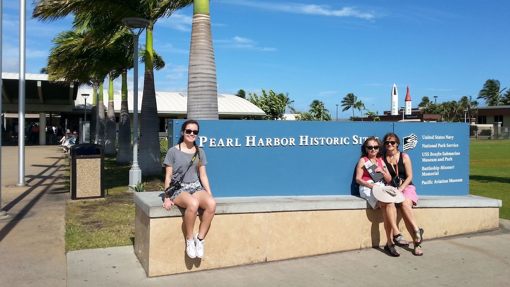Three women sit next to Pearl Harbor Historic Site sign in Oahu