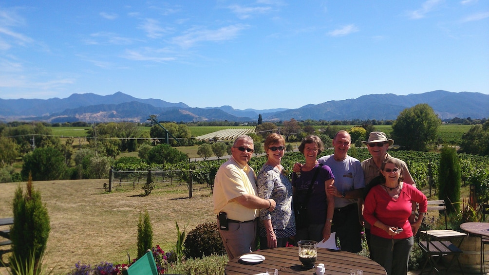 Show item 4 of 5. Group posing at outdoor patio with beautiful vineyards in the distance.