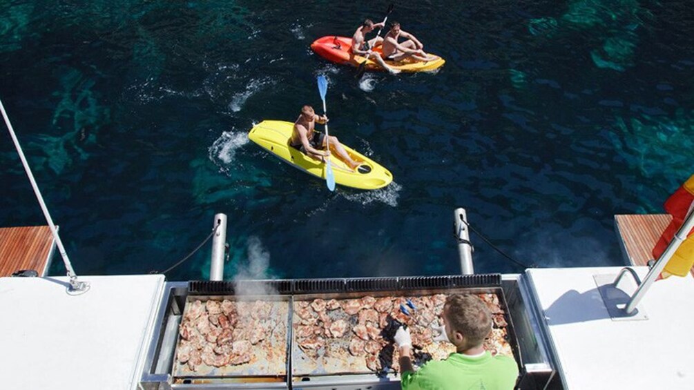 Man at a grill looks down at Kayakers next to a Catamaran