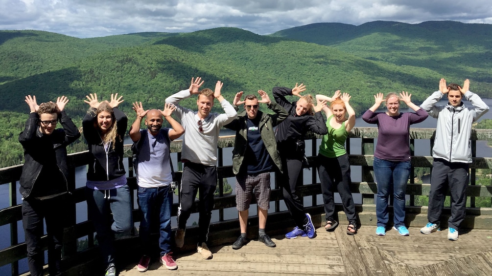 Show item 1 of 4. A tour group making moose antlers with their hands in a scenic view point of Canadian hills