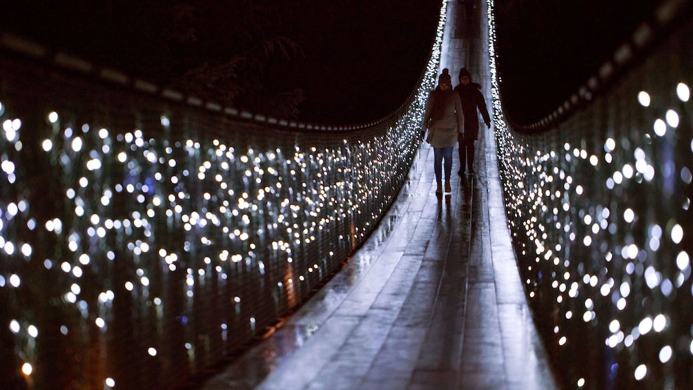 People walking across Capilano Bridge lit up with holiday lights at night in Vancouver