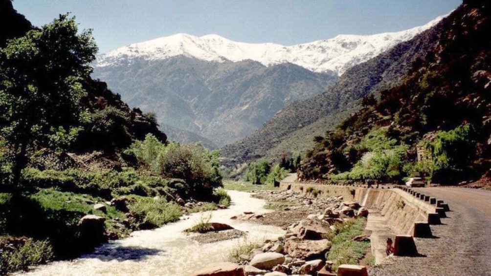 Dirt road and greenery bordering river with snow capped mountains in background in Ourika Valley