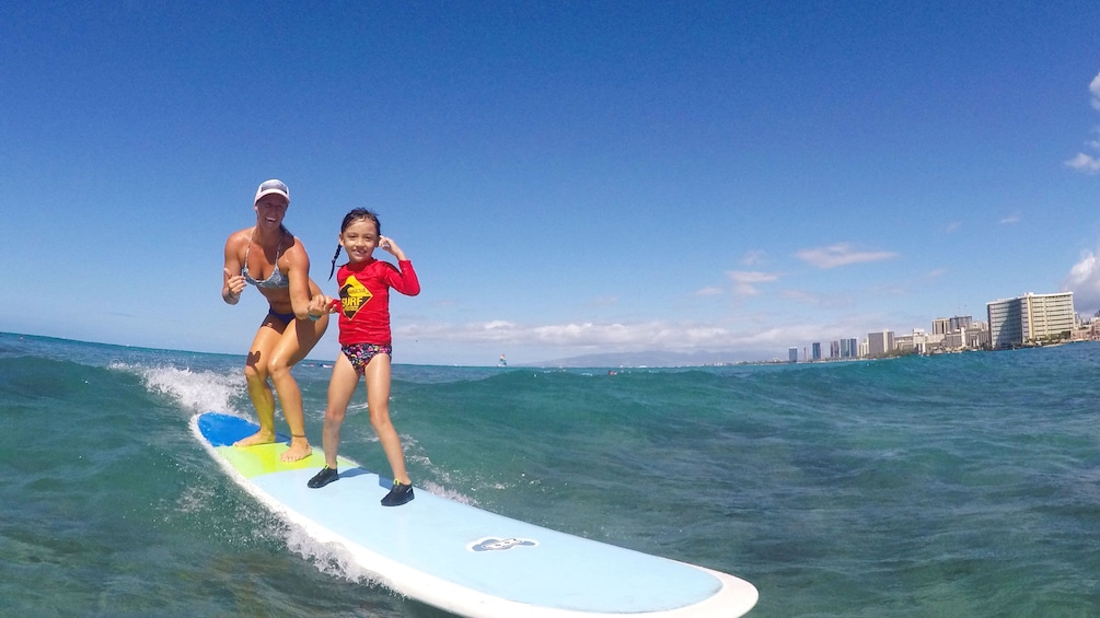Instructor holds small girl's hand as they ride wave on surfboard in bay in Oahu