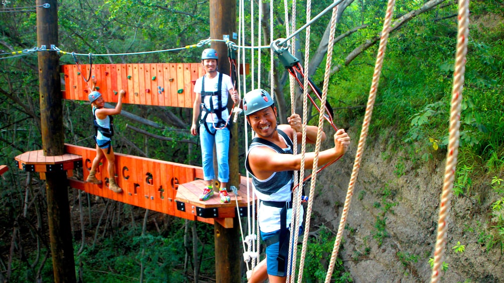 Three people climbing a rope bridge and peg wall suspended in the air.