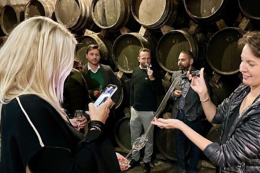 Aged brandy is extracted directly from the cask and served into the glass of the visitors to taste exclusive spirits. A group of women and men tasting liquors inside the cellar, and historic cave. 