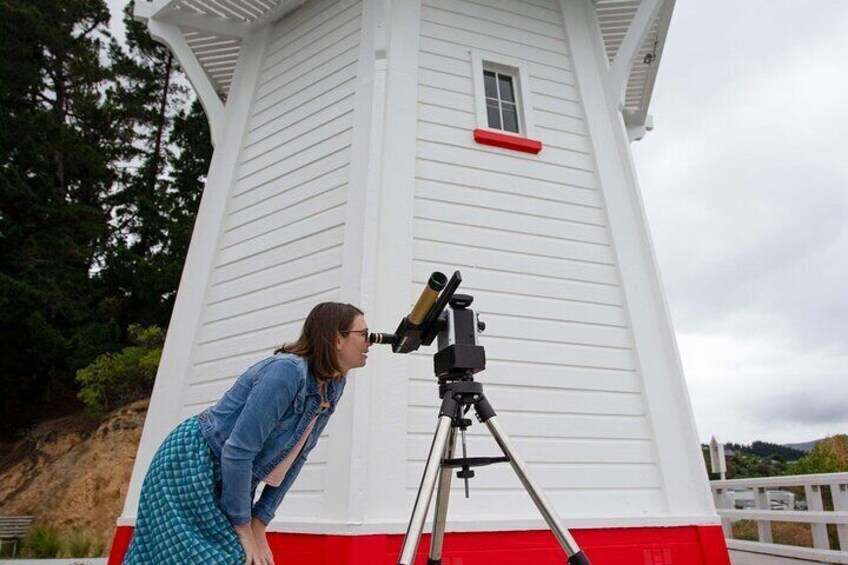 Lost in the brilliance of the sun's dance, a woman gazes through a solarscope beside the iconic Akaroa Lighthouse. Join us at this historic landmark for a Solar Explorer experience like no other. 